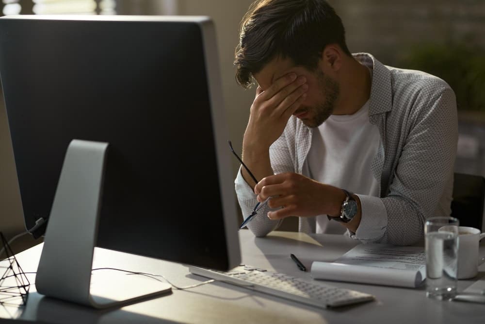 Overworking businessman in the office who experiences a throbbing headache and extreme exhaustion while laboring on his desktop computer